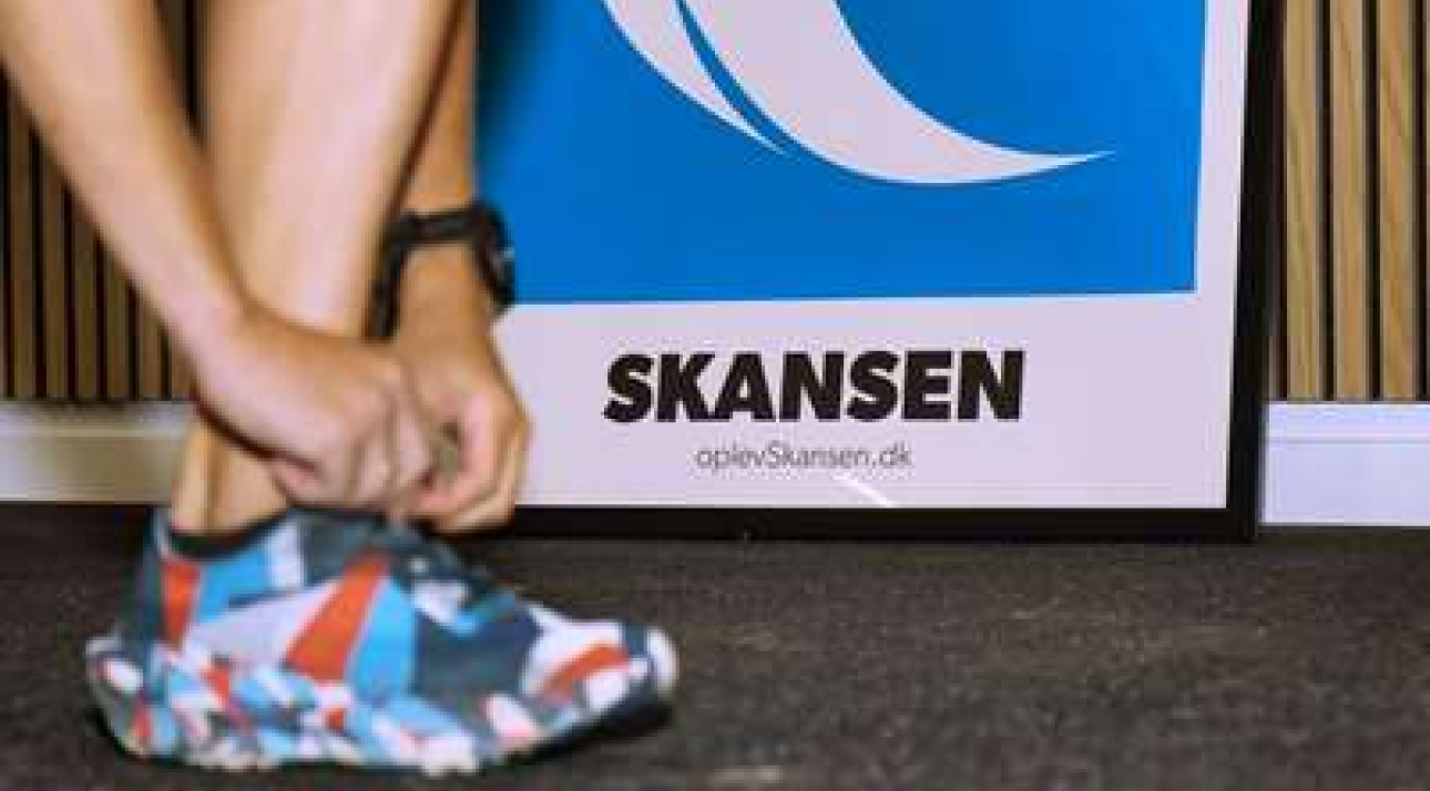 Person binder snørrebånd med plakat af Skansen i baggrunden